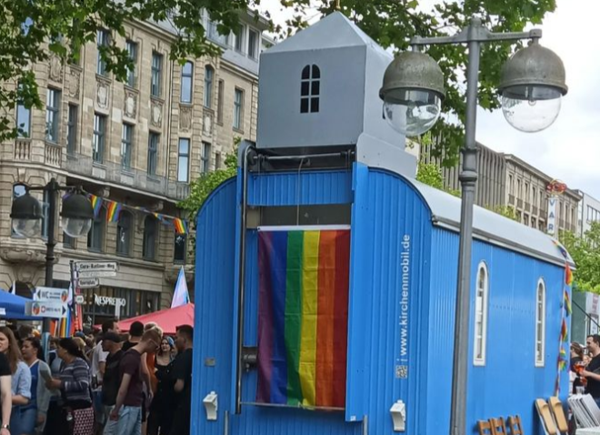 Blaue Bauwagenkirche in einer Menschenmenge. Am Turm hängt eine Regenbogenfahne